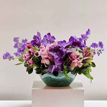Bouquet of purple and pink flowers in a textured green vase on a white surface with a light gray background