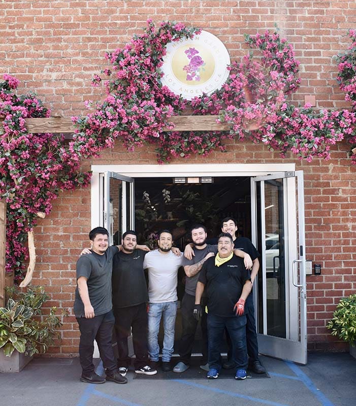 Group of people posing in front of a brick building with floral decorations and a logo.