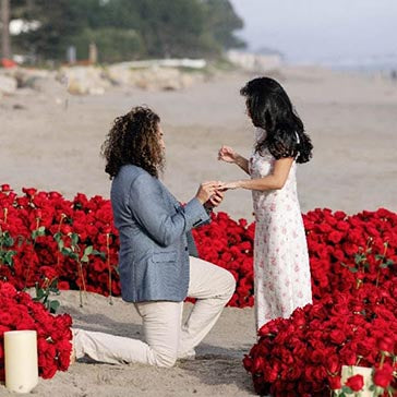 Man proposing to a woman on a beach with red flowers and candles.
