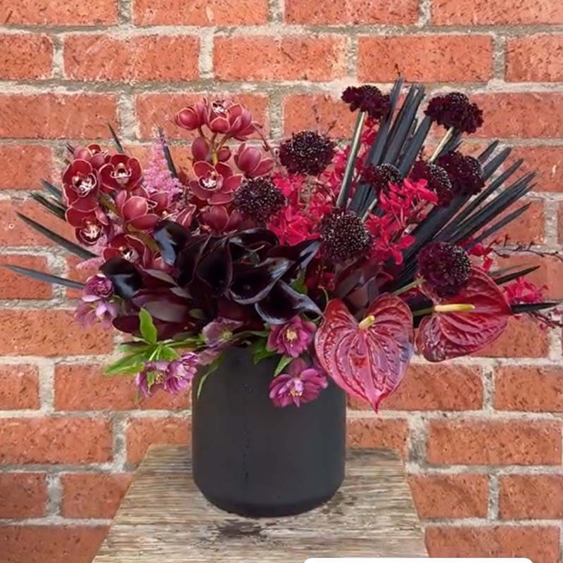 Bouquet of dark pink and purple flowers in a black vase against a brick wall.