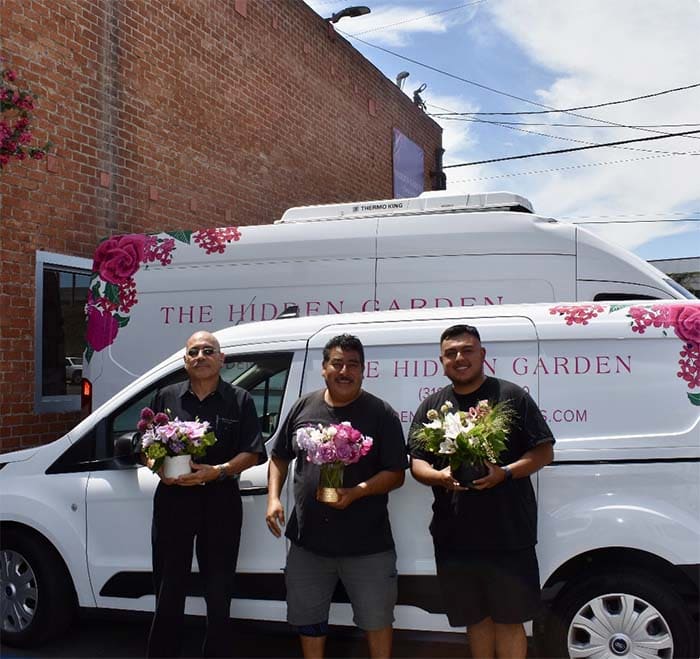 Three men holding flowers in front of a van with 'The Hidden Garden' branding.