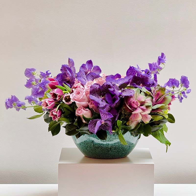 Bouquet of purple and pink flowers in a green vase on a white pedestal against a light gray background
