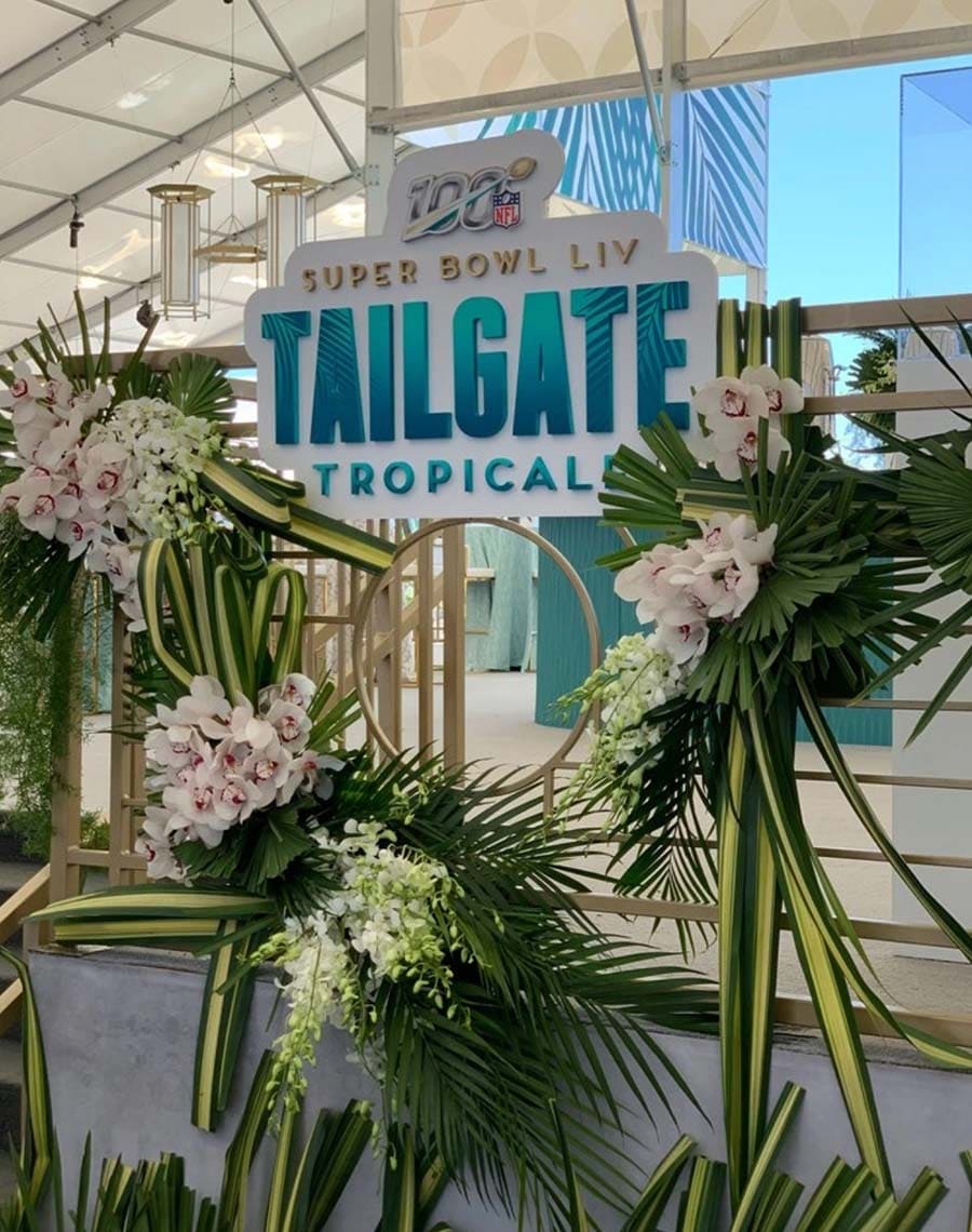 Decorative setup with 'Super Bowl LIV Tailgate Tropical' sign and floral arrangements.