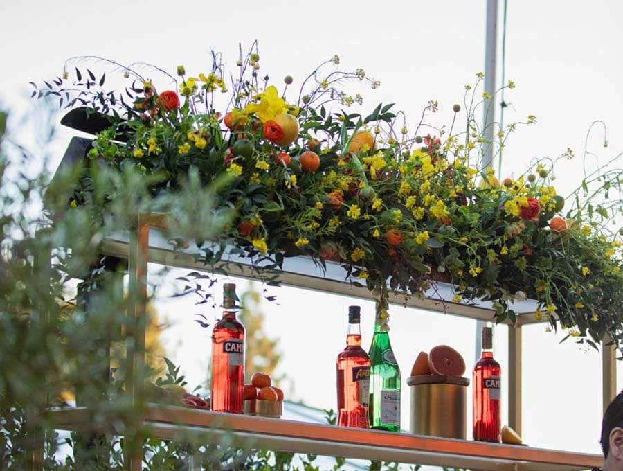 Decorative floral arrangement above a bar with bottles and fruit on a sunny day.