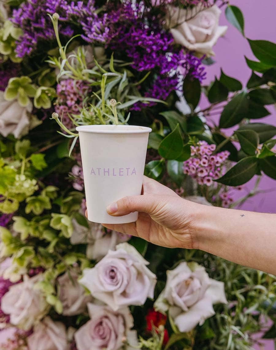 Hand holding a white cup with 'ATHLETA' branding in front of a floral arrangement.