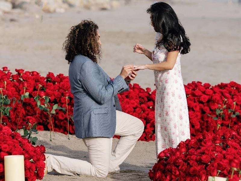Man proposing to a woman in a field of red flowers on a beach.