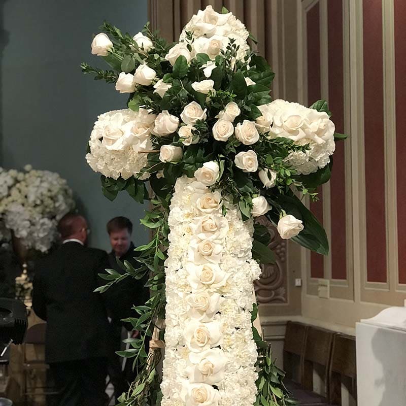 Floral arrangement in the shape of a cross with white flowers and green leaves, set against a decorative wall.
