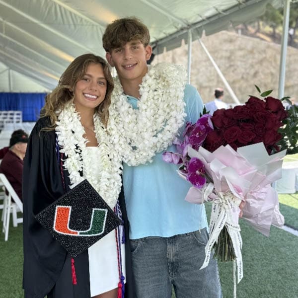Two people celebrating a graduation ceremony with flowers and a cap, standing under a tent.