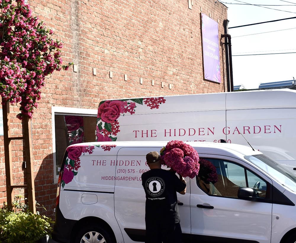 White van with 'The Hidden Garden' branding, person holding flowers, brick building in background