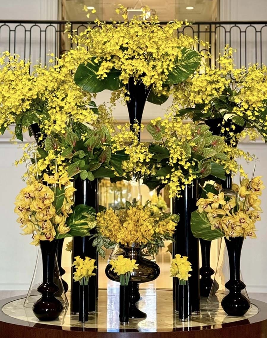 Decorative floral arrangement with yellow flowers and green leaves in black vases on a reflective surface.