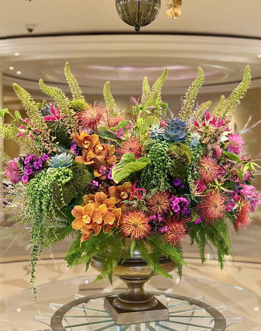 Colorful floral arrangement with various plants in a decorative urn on a reflective surface.