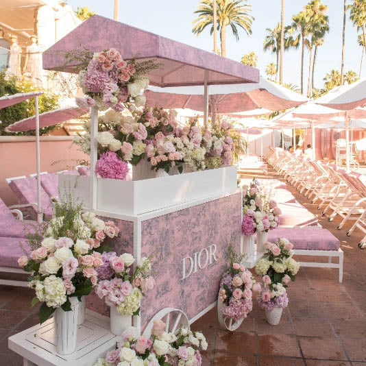 Decorative cart with 'Dior' branding surrounded by floral arrangements and pink chairs under umbrellas.