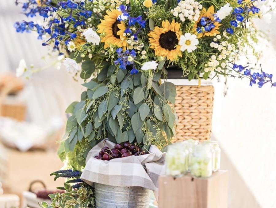 Decorative floral arrangement with sunflowers, blue flowers, and greenery in a wicker basket.