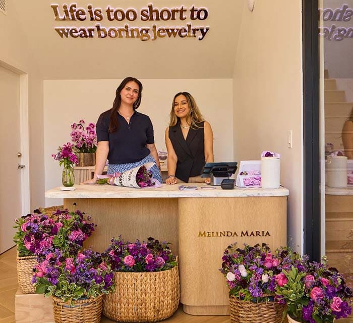 Two women standing behind a counter with 'Melinda Maria' branding, surrounded by floral arrangements.