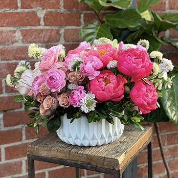Bouquet of pink and white flowers in a white vase on a wooden stool against a brick wall.