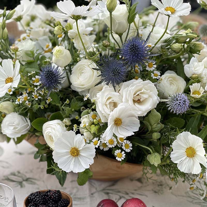 Bouquet of white flowers with greenery and blue thistle in a wooden basket.