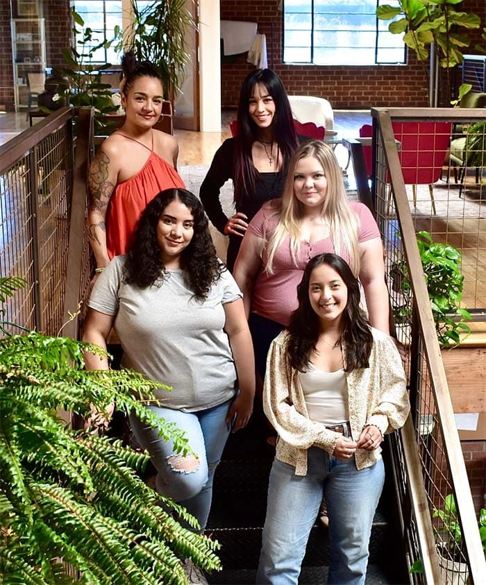 Five women posing together in a modern indoor setting with plants and furniture.
