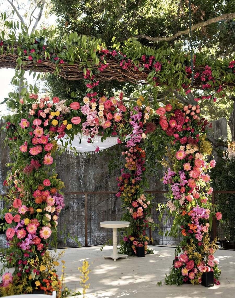 Floral archway decorated with pink, red, and yellow flowers in an outdoor setting.