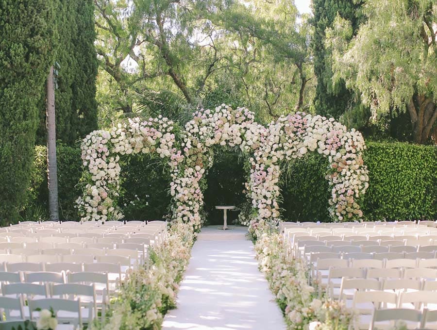 Floral archway at a wedding ceremony with chairs and a table in a garden setting.