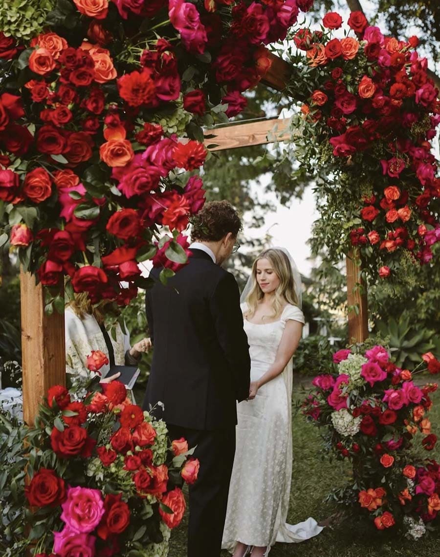 Couple standing under a floral archway with red and pink flowers.