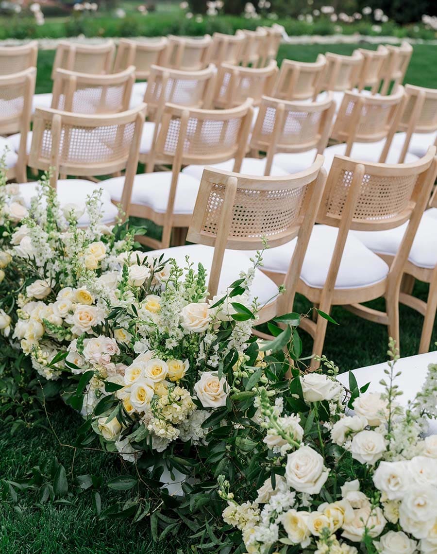 Decorative floral arrangement with white and yellow flowers in front of rows of beige chairs with white cushions.