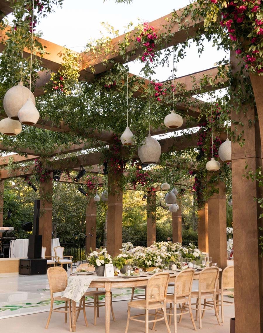 Outdoor dining setup under a wooden pergola with hanging lights and floral decorations.