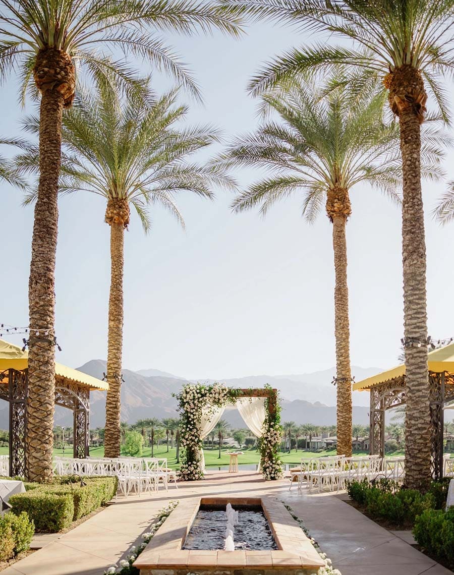 Outdoor wedding setup with palm trees, arch, and mountains in the background
