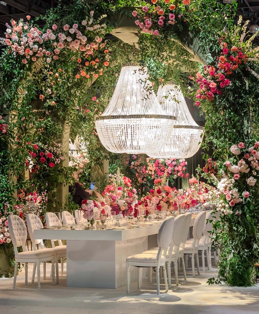 Dining table set with floral decorations and a chandelier in a lush green and floral setting.