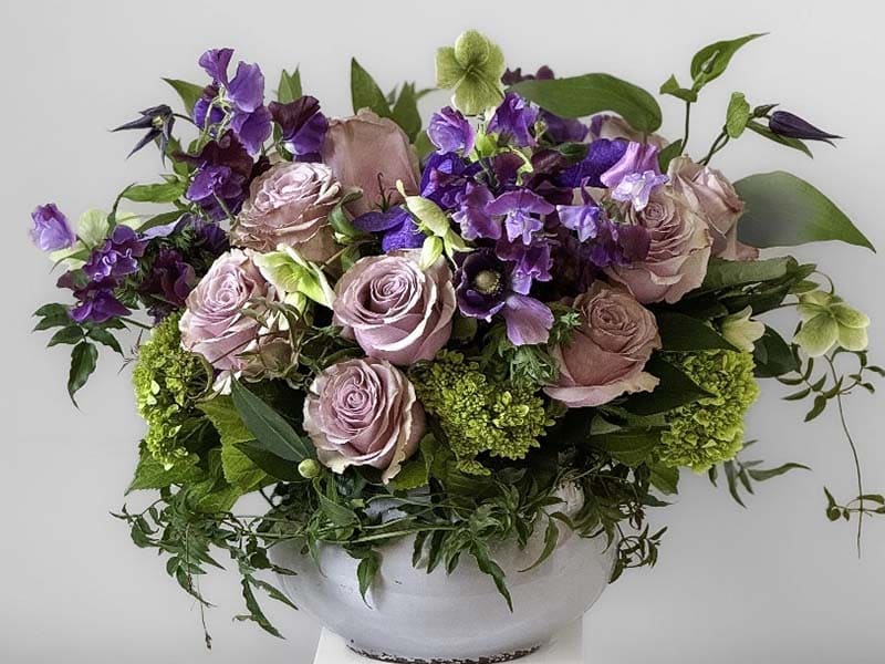 Floral arrangement with purple and pink roses, greenery, and a white vase on a light background